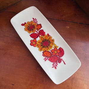 May include: A rectangular white ceramic serving platter with a floral design. The flowers are in shades of orange, yellow, and red, with red leaves and stems. The platter is a decorative piece for serving food.
