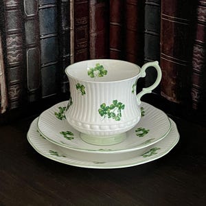 May include: A white teacup, saucer, and plate set with a green shamrock design. The teacup has a ribbed texture and a green handle. The set is displayed on a dark surface against a backdrop of antique books.