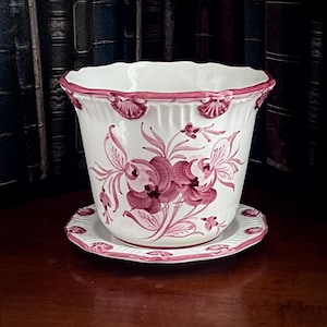 May include: White ceramic plant pot with a scalloped rim and matching saucer. The pot features a floral design in shades of pink and a pink rim. The pot is sitting on a wooden surface.