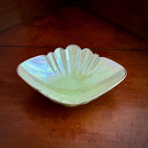 May include: A shallow, shell-shaped dish with a pale, iridescent yellow finish and a metallic silver-toned rim. The bowl features a scalloped edge and a ribbed, fan-like texture radiating from the centre, creating a reflective, pearlescent surface.