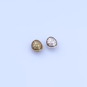 Pode incluir: Duas pequenas contas de metal em forma oval. Uma é dourada, a outra prateada. Ambas as contas apresentam um padrão geométrico em relevo que se assemelha a uma carapaça de tartaruga. As contas são exibidas em um fundo azul claro.