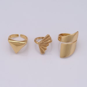 Puede incluir: Tres anillos dorados con diferentes diseños geométricos. El primer anillo es de forma triangular, el segundo anillo es de forma de abanico y el tercer anillo es de forma rectangular curvada.