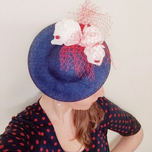 May include: A blue straw hat with a red and white flower embellishment. The hat is decorated with a red mesh veil and white flowers with red centers.