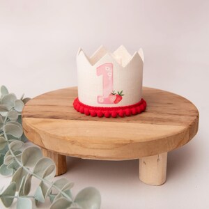 May include: A white fabric crown with a pink number 1 and strawberry design, trimmed with red pom-poms. The crown sits on a small, round wooden stand. The stand is approximately 6 inches in diameter.