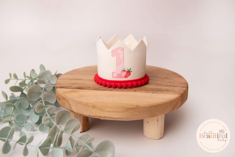 May include: A white fabric crown with a pink number 1 and strawberry design, trimmed with red pom-poms. The crown sits on a small, round wooden stand. The stand is approximately 6 inches in diameter.