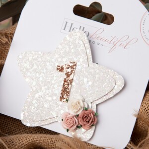 Pu&ograve; includere: Stella bianca con glitter con un numero uno in oro rosa e fiori rosa. La stella &egrave; attaccata a una carta bianca con il testo "Hello beautiful bows".