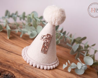 Peut inclure: Un chapeau de fête conique en feutre blanc avec un pompon blanc moelleux sur le dessus. Le chapeau a un numéro "1" en paillettes or rose sur le devant.