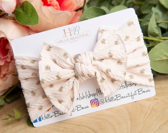 May include: A cream-colored fabric headband with a large bow tied in the center. The bow is decorated with a small floral pattern in shades of yellow, brown, and blue. The headband is displayed on a white card with the text "Hello Beautiful Bows" and a social media handle.