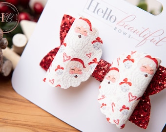 May include: Two Christmas-themed hair bows. The bows feature a white background with Santa Claus faces, candy canes, and bells. The center and accents are red glitter. The bows are on a white card with the text "Hello Beautiful Bows".