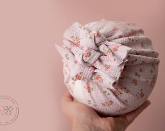 May include: A pink floral fabric turban headband with a knotted bow on top. The headband is made of a soft, stretchy fabric and is perfect for newborns and babies.