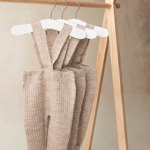 May include: Three pairs of beige ribbed knit baby overalls hanging on white wooden hangers on a wooden clothing rack.