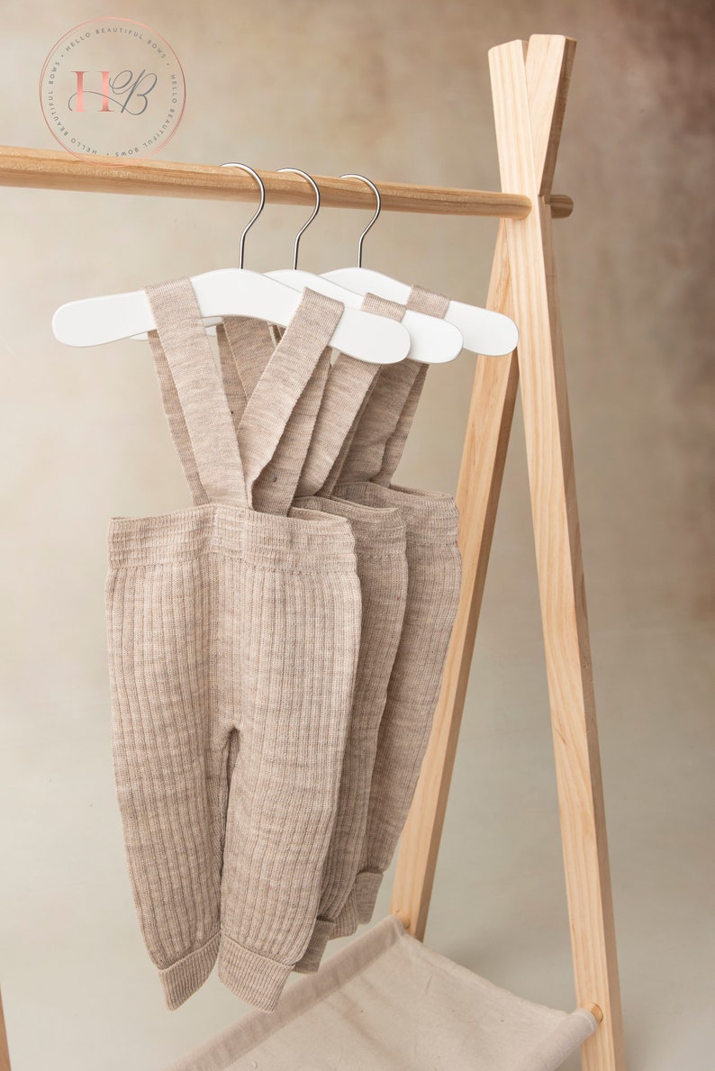 May include: Three pairs of beige ribbed knit baby overalls hanging on white wooden hangers on a wooden clothing rack.
