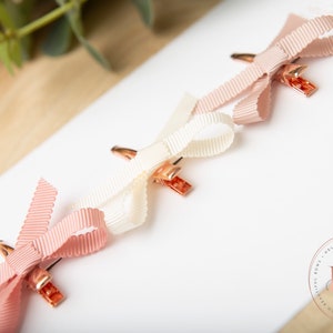 May include: Three small hair clips with bows. The bows are made of pink and white ribbon. The clips are gold colored.