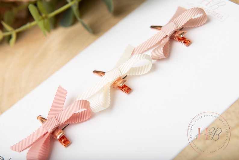 May include: Three small hair clips with bows. The bows are made of pink and white ribbon. The clips are gold colored.