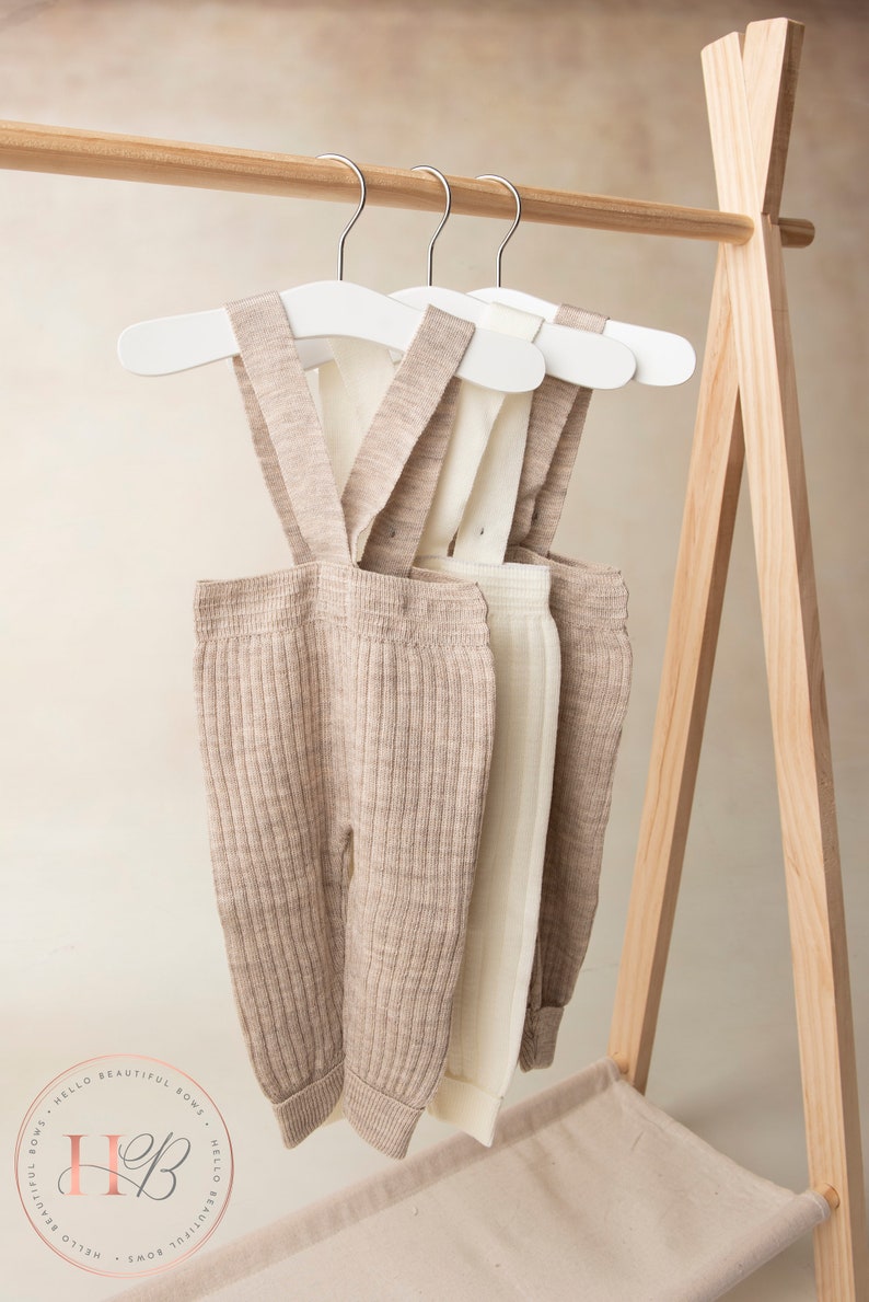May include: Three pairs of beige and white ribbed knit baby overalls hanging on wooden hangers on a wooden rack. The overalls have adjustable straps.