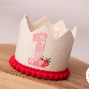 May include: A cream-colored fabric crown with a red pom-pom trim. The crown features a pink number 1 with daisy accents and a strawberry illustration. The crown is designed for a baby's first birthday.