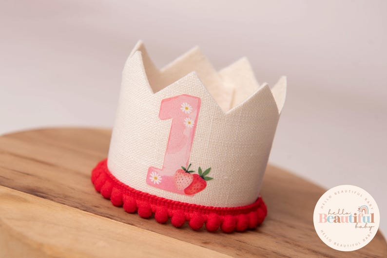 May include: A cream-colored fabric crown with a red pom-pom trim. The crown features a pink number 1 with daisy accents and a strawberry illustration. The crown is designed for a baby's first birthday.