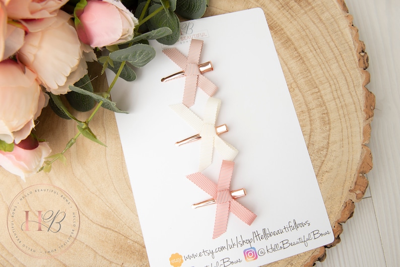 May include: Three pink, white, and blush satin ribbon hair bows with gold clips. The bows are arranged vertically on a white card with the text "www.etsy.com/uk/shop/HelloBeautifulBows @HelloBeautifulBows"