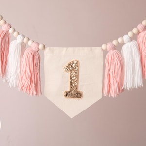 Peut inclure: Guirlande de fanions avec un num&eacute;ro 1 dor&eacute; &agrave; paillettes sur un fanion cr&egrave;me. La guirlande est orn&eacute;e de pompons en fil rose et blanc et de perles en bois. Id&eacute;ale pour une f&ecirc;te de premier anniversaire.