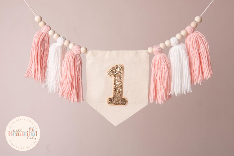 Peut inclure: Guirlande de fanions avec un num&eacute;ro 1 dor&eacute; &agrave; paillettes sur un fanion cr&egrave;me. La guirlande est orn&eacute;e de pompons en fil rose et blanc et de perles en bois. Id&eacute;ale pour une f&ecirc;te de premier anniversaire.