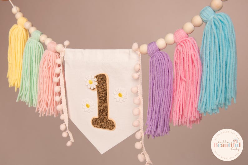 Op de afbeelding: Een pastelkleurige slinger met een groot cijfer 1, madeliefjesaccenten en kleurrijke garentoefjes. De slinger is geregen met houten kralen en pompom-afwerking, ideaal voor een eerste verjaardag.