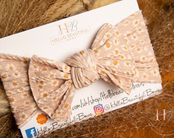 May include: A pink and white knotted headband with a daisy print. The headband is on a white card with the text "Hello Beautiful Bows" and social media handles.