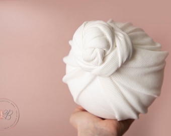 May include: A white, ribbed knit baby turban with a knotted top. The soft fabric is wrapped around a round shape, creating a cozy and stylish accessory. The background is a soft pink.