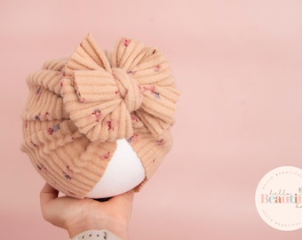 May include: A beige turban with a floral pattern and a large bow. The turban is made of a soft, stretchy fabric and is perfect for newborns.
