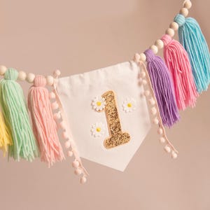 Op de afbeelding: Een kleurrijke banner met een witte wimpel met een gouden glitter nummer 1 en madeliefjesaccenten. De banner is versierd met kwasten in geel, mintgroen, roze, paars en blauw, en houten kralen. Perfect voor een eerste verjaardag.