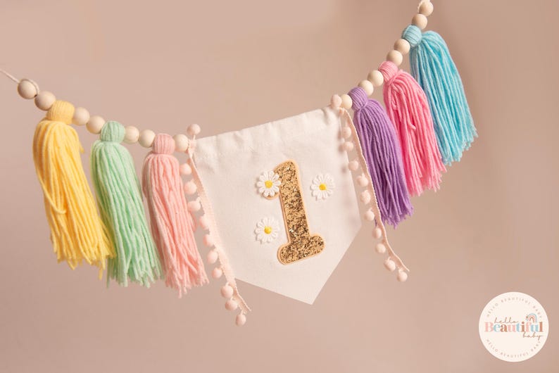 Op de afbeelding: Een kleurrijke banner met een witte wimpel met een gouden glitter nummer 1 en madeliefjesaccenten. De banner is versierd met kwasten in geel, mintgroen, roze, paars en blauw, en houten kralen. Perfect voor een eerste verjaardag.
