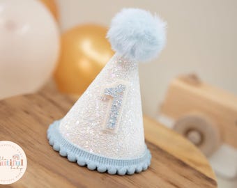 May include: A white and blue glitter party hat with a blue pom-pom on top and a number one on the front. The hat is sitting on a wooden surface.