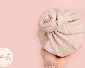 May include: A beige ribbed knotted turban headband for babies. The headband is made of soft, stretchy fabric and is perfect for newborns and infants.
