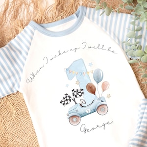 May include: A white baby outfit with blue striped sleeves and pants. The front of the shirt features a watercolor illustration of a blue race car with the number 1, balloons, and the text "When I wake up I will be" and "George".
