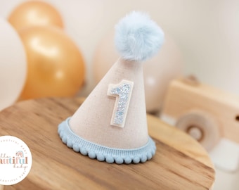 May include: A light blue and white felt party hat with a fluffy white pom pom on top. The hat has a silver glitter number one on the front.