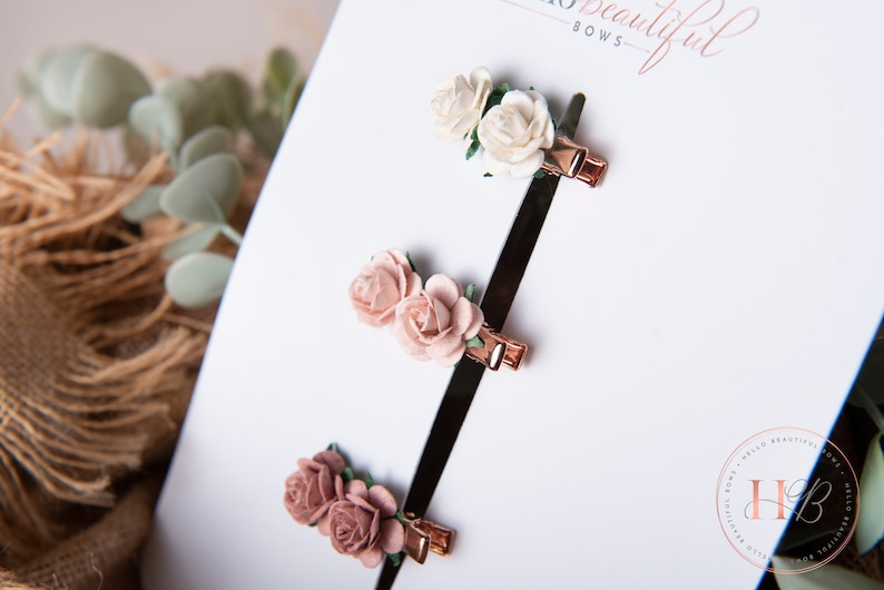 May include: Three sets of hair clips, each featuring two small fabric roses. The roses are in shades of pink and white. The clips are gold coloured.