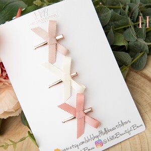 May include: Set of three hair clips with pink, white, and blush bows. The clips have a rose gold finish. The clips are displayed on a white card with the text "Hello Beautiful Bows" and a website address.