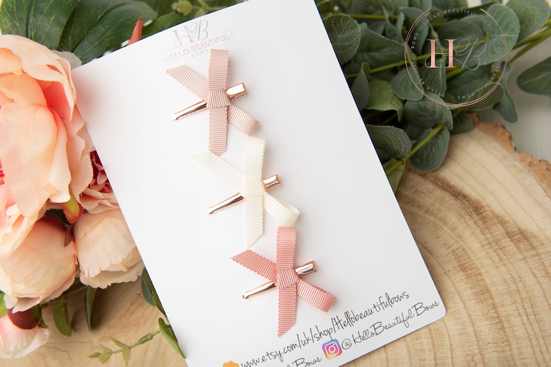 May include: Set of three hair clips with pink, white, and blush bows. The clips have a rose gold finish. The clips are displayed on a white card with the text "Hello Beautiful Bows" and a website address.
