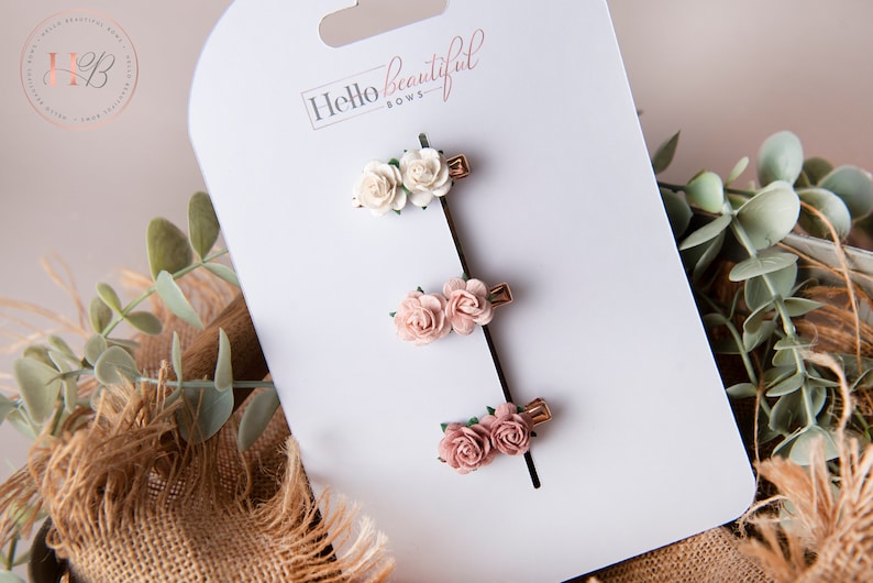 May include: Three small hair clips with two white and two pink fabric flowers on each clip. The clips are on a white card with the text "Hello Beautiful Bows".
