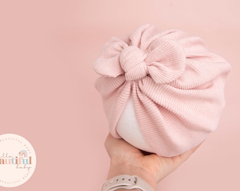 May include: A pink turban with a large bow on top. The turban is made of a soft, ribbed fabric.