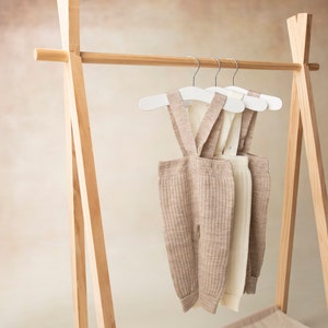 May include: Three pairs of beige and white ribbed knit baby overalls hanging on white hangers on a wooden clothing rack.