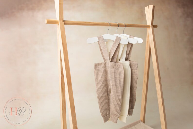 May include: Three pairs of beige and white ribbed knit baby overalls hanging on white hangers on a wooden clothing rack.