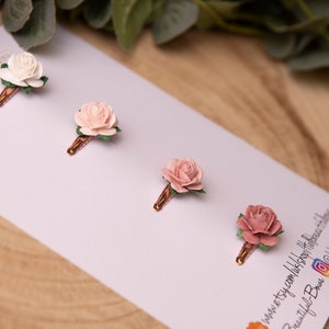May include: Four small flower hair clips with a rose design. The clips are arranged in a row on a white background. The flowers are in shades of pink and white.