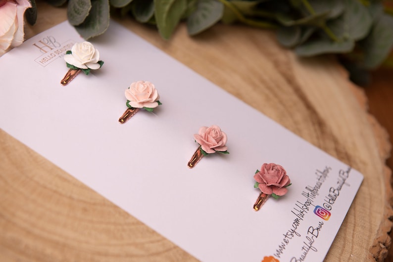 May include: Four small flower hair clips with a rose design. The clips are arranged in a row on a white background. The flowers are in shades of pink and white.
