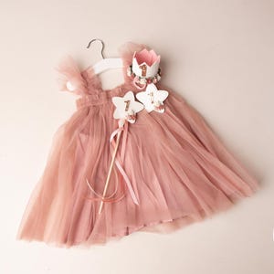 Op de afbeelding: Stofroze tule jurk met bijpassende kroon en sterrenstaven. De jurk hangt aan een witte hanger. De kroon en de staven hebben bloemaccenten en het nummer "1". De jurk is voor een baby.