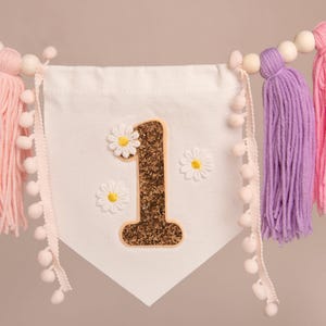 Op de afbeelding: Een witte slinger met een gouden glitter "1" en madeliefjes. De slinger heeft gekleurde kwasten in mintgroen, roze, paars en blauw. Houten kralen en pompom-randen maken het ontwerp af.