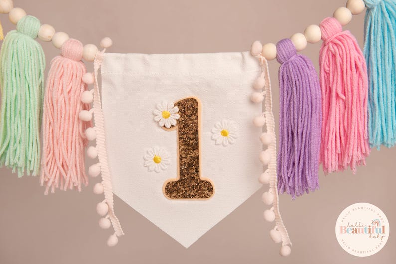 Op de afbeelding: Een witte slinger met een gouden glitter "1" en madeliefjes. De slinger heeft gekleurde kwasten in mintgroen, roze, paars en blauw. Houten kralen en pompom-randen maken het ontwerp af.