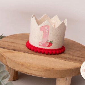 May include: A linen crown with a red pom-pom trim, featuring a pink number 1 and strawberry graphic. The crown sits on a wooden stand. The crown is designed for a first birthday celebration.