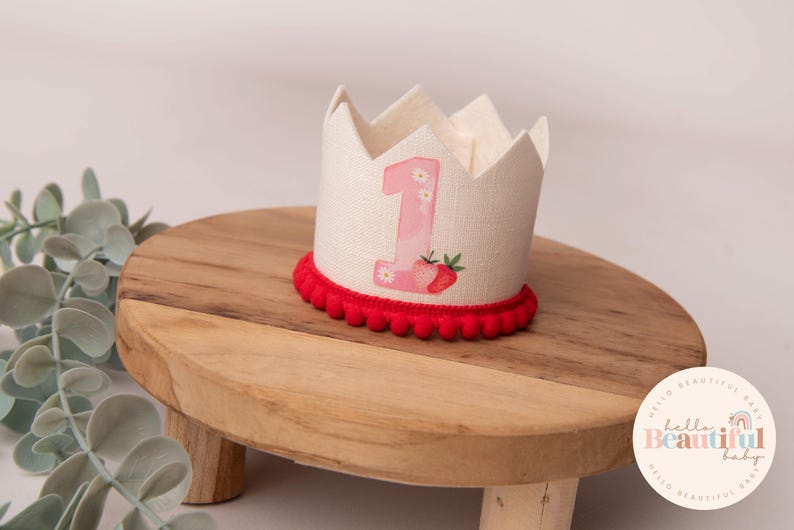 May include: A linen crown with a red pom-pom trim, featuring a pink number 1 and strawberry graphic. The crown sits on a wooden stand. The crown is designed for a first birthday celebration.