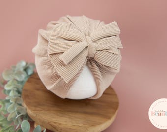 May include: A beige turban with a large bow on top. The turban is made of a soft, stretchy fabric and is perfect for newborn photography props.