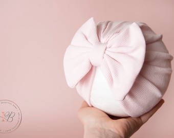 May include: A pink ribbed turban with a large bow on top. The turban is made of soft, stretchy fabric and is perfect for newborns.